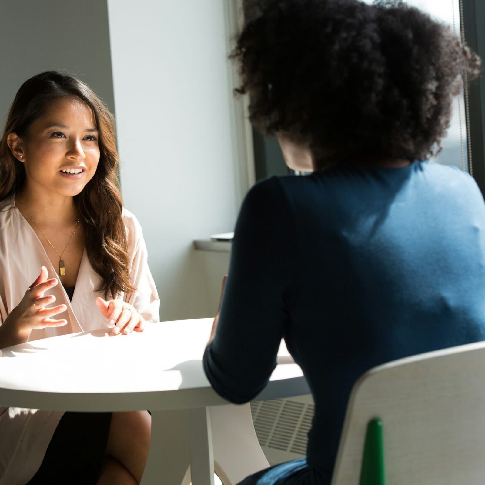 01-icf-guide-experience-coaching-differences Two woman sit at a table in a semi professional setting talking to each other.