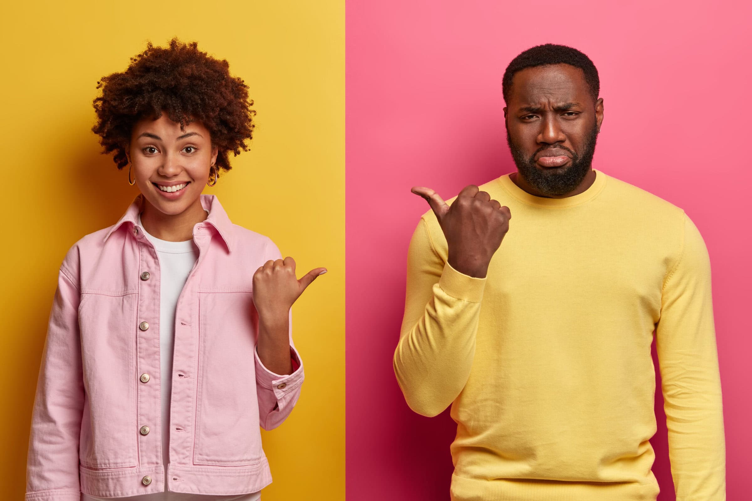 Studio shot of cheerful black woman and gloomy bearded man point thumbs at each other, blame and express different emotions, dressed casually, pose over bright two colored wall. Its he or she A woman and a man pointing their thumbs at each other, the woman is smiling, while the man is pouting.