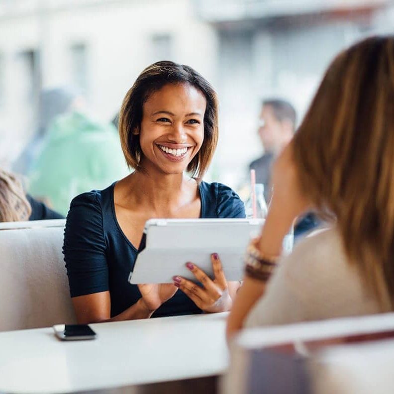 Coaching Conversation in Cafe A smiling woman holding a tablet, chatting with another woman at a cafe.