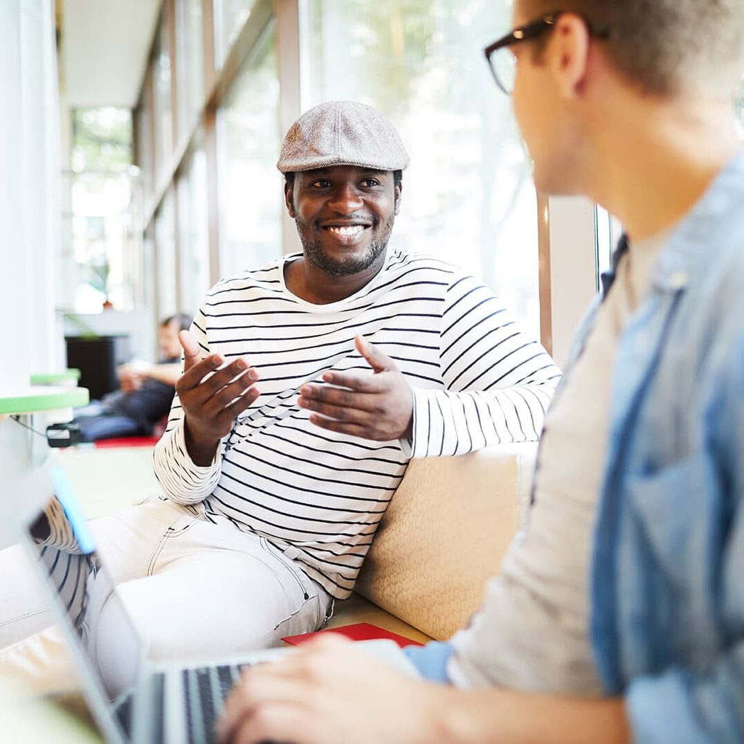icf-become-a-coach-community-events-two-men-connection-discussion Two men engaged in a lively conversation at a community event, one smiling warmly and gesturing expressively, seated indoors near large windows with natural light filtering in.