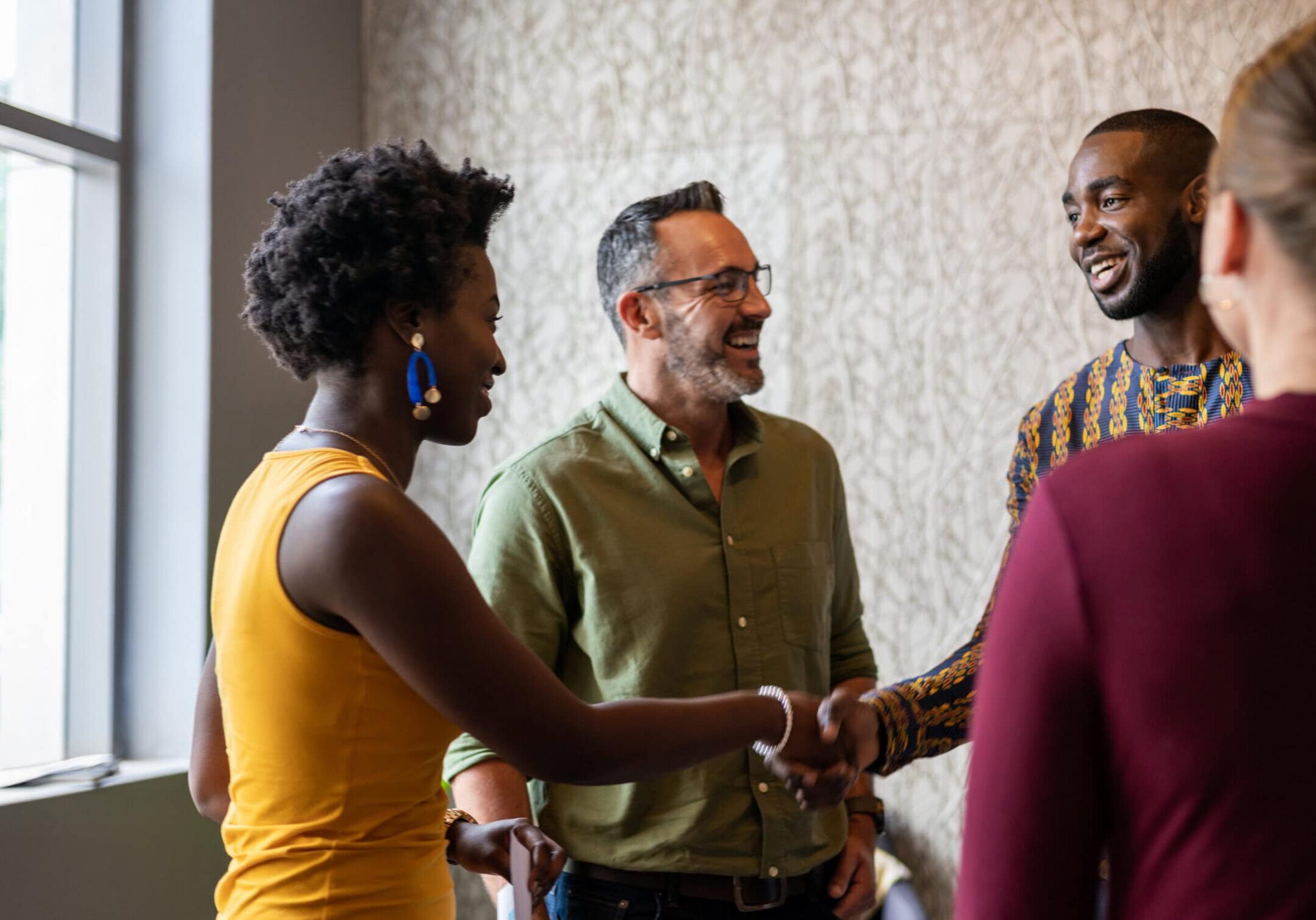 Diversity in Coaching Connections A diverse group of coaches connect warmly, shaking hands and smiling in an office setting. Two individuals greet each other in the foreground, while others stand nearby, fostering a friendly and collaborative atmosphere.