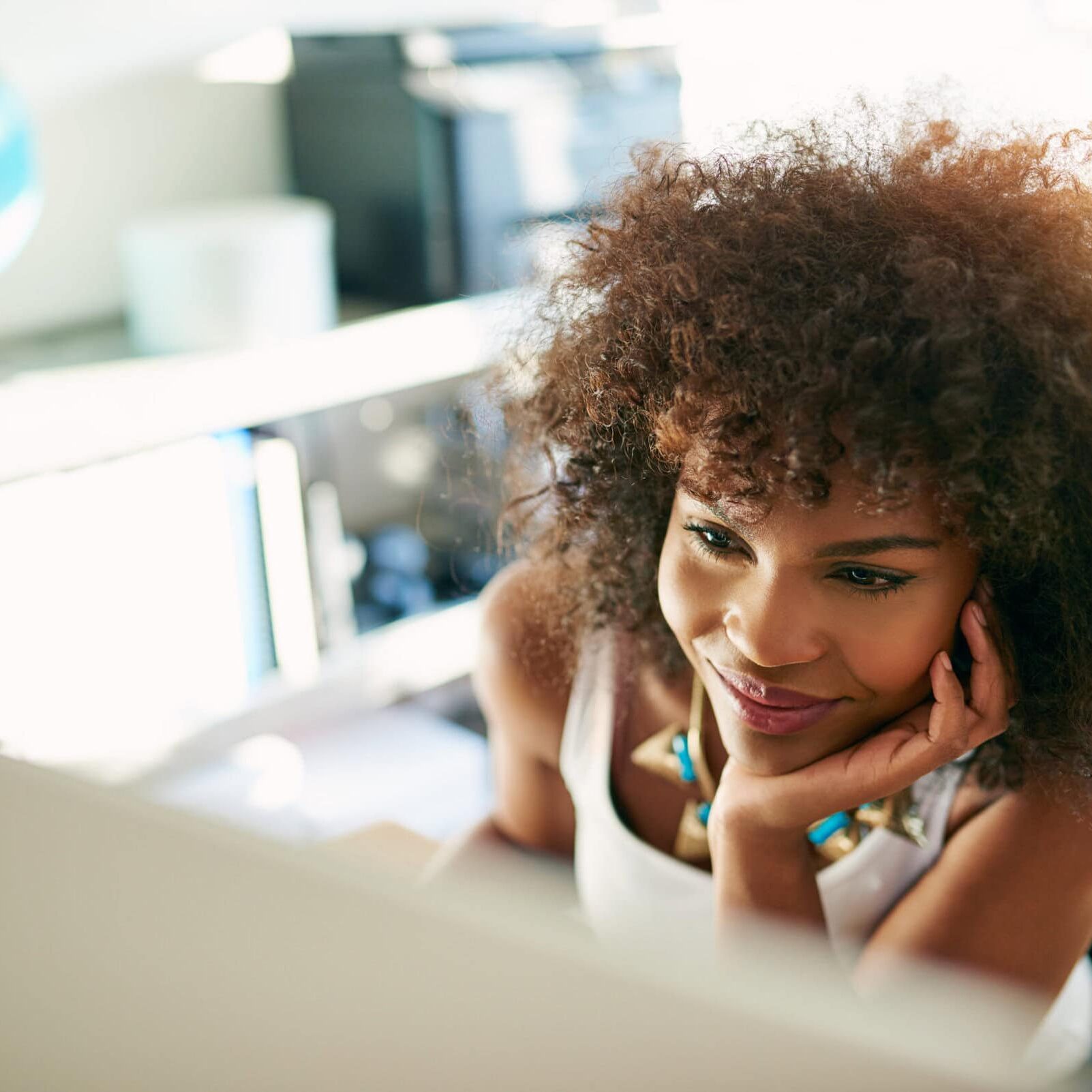Coach Professional Development A young woman with curly hair smiling while working on coach professional development at a desk, with a globe and computer visible in a bright office setting.