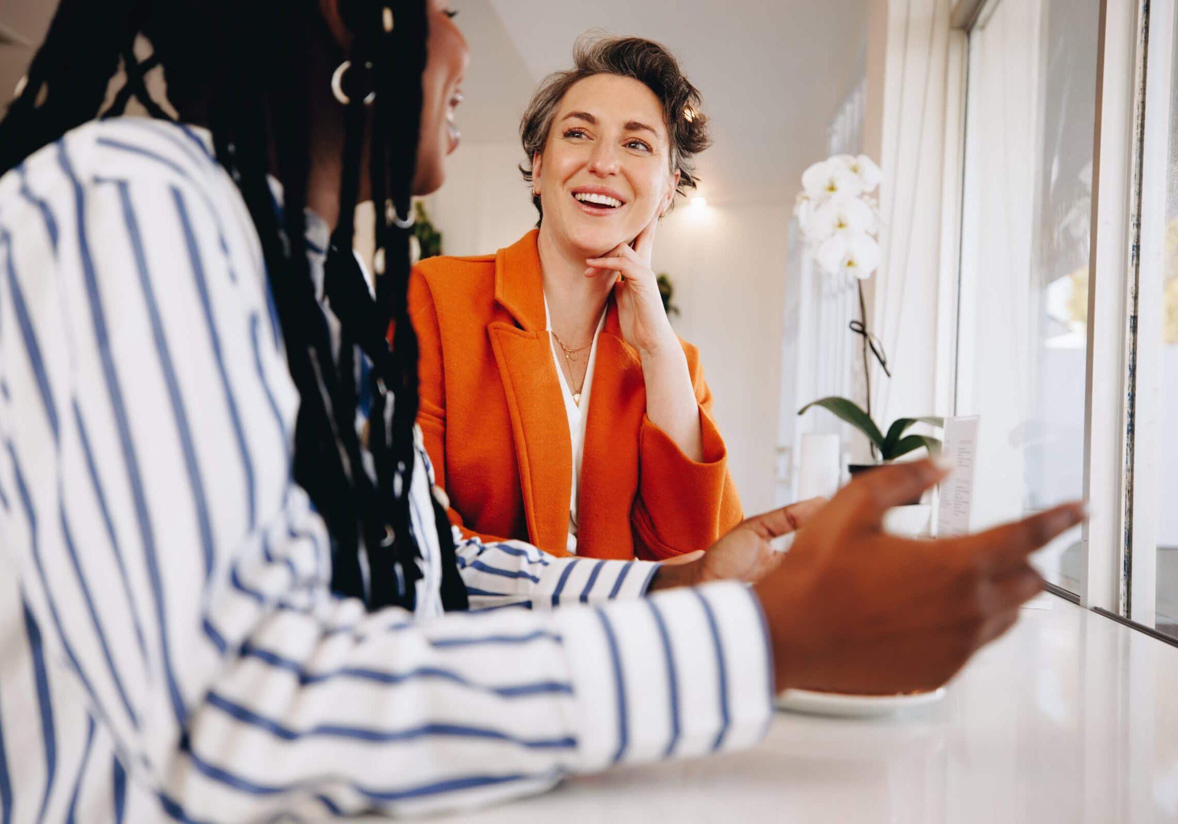 Coaching Interaction A smiling woman in an orange blazer having a friendly coaching interaction with another woman in a cozy indoor setting.