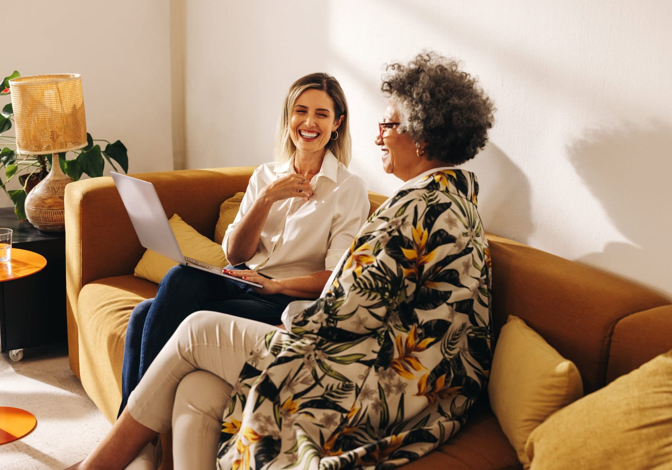 Friendly Coaching Session Two women engaged in a friendly coaching session on a couch, with one holding a laptop, in a cozy, sunlit room.