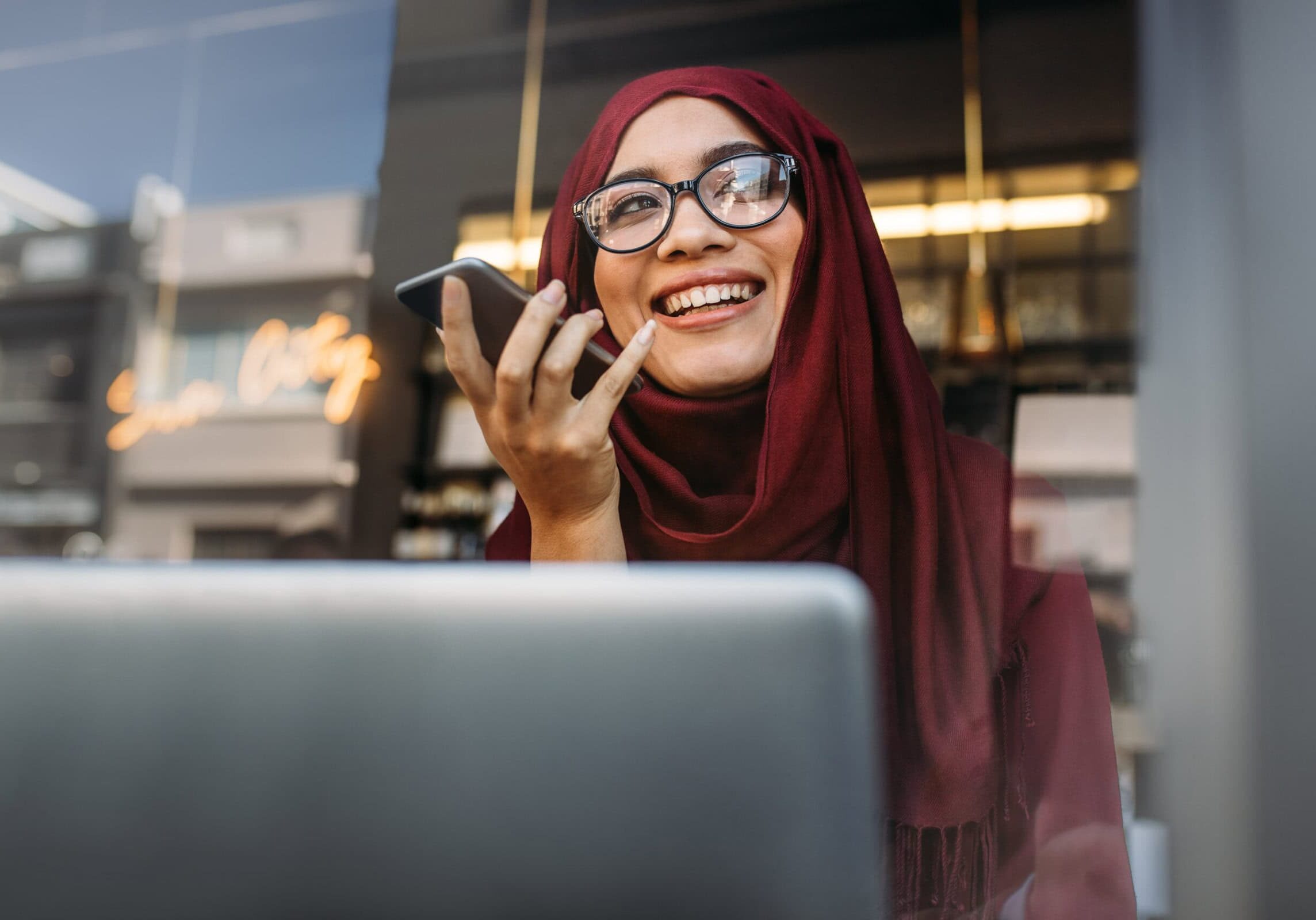 Inclusive Coaching Session Young professional woman in a maroon headscarf smiling while having a coaching session on her phone in front of a laptop, symbolizing connection and inclusivity within the ICF community.
