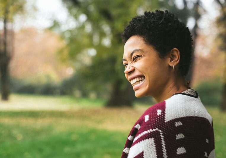 icf-join-icf-joy-from-coaching A smiling woman with short curly hair wearing a colorful patterned jacket in a natural outdoor setting, portraying positivity and joy from the coaching profession.