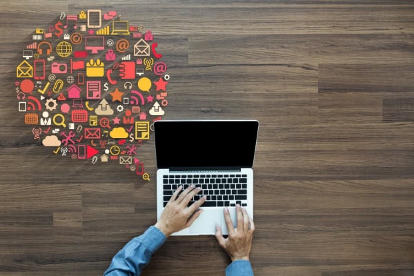 Person working at a computer with a large thought bubble to the left of the laptop comprised of many small icons typically associated with computers in varying colors