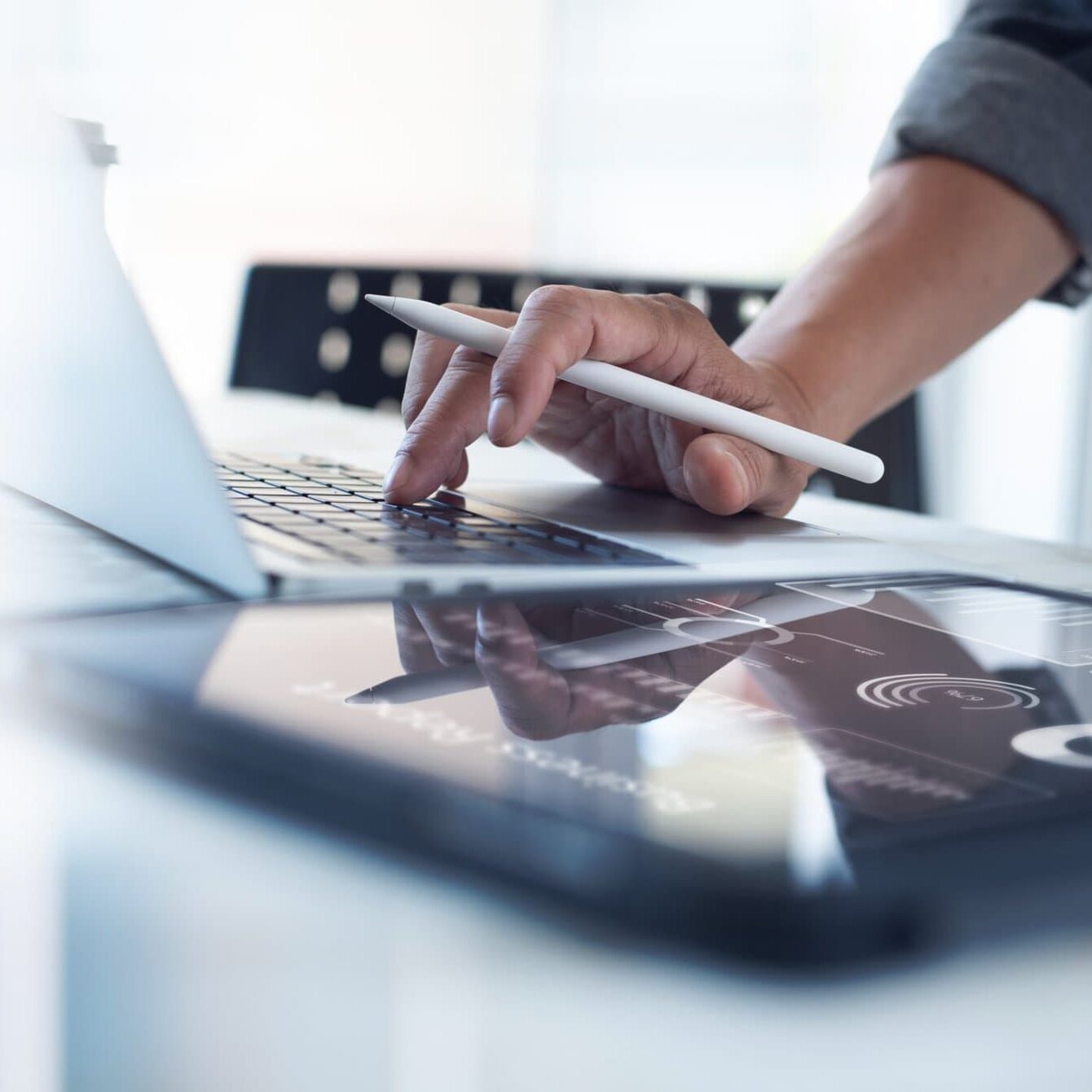 Businessman,Working,On,Laptop,,Using,Mobile,Phone,At,Modern,Office, person with one hand on a laptop keyboard with an apple pencil and their other hand on an ipad screen
