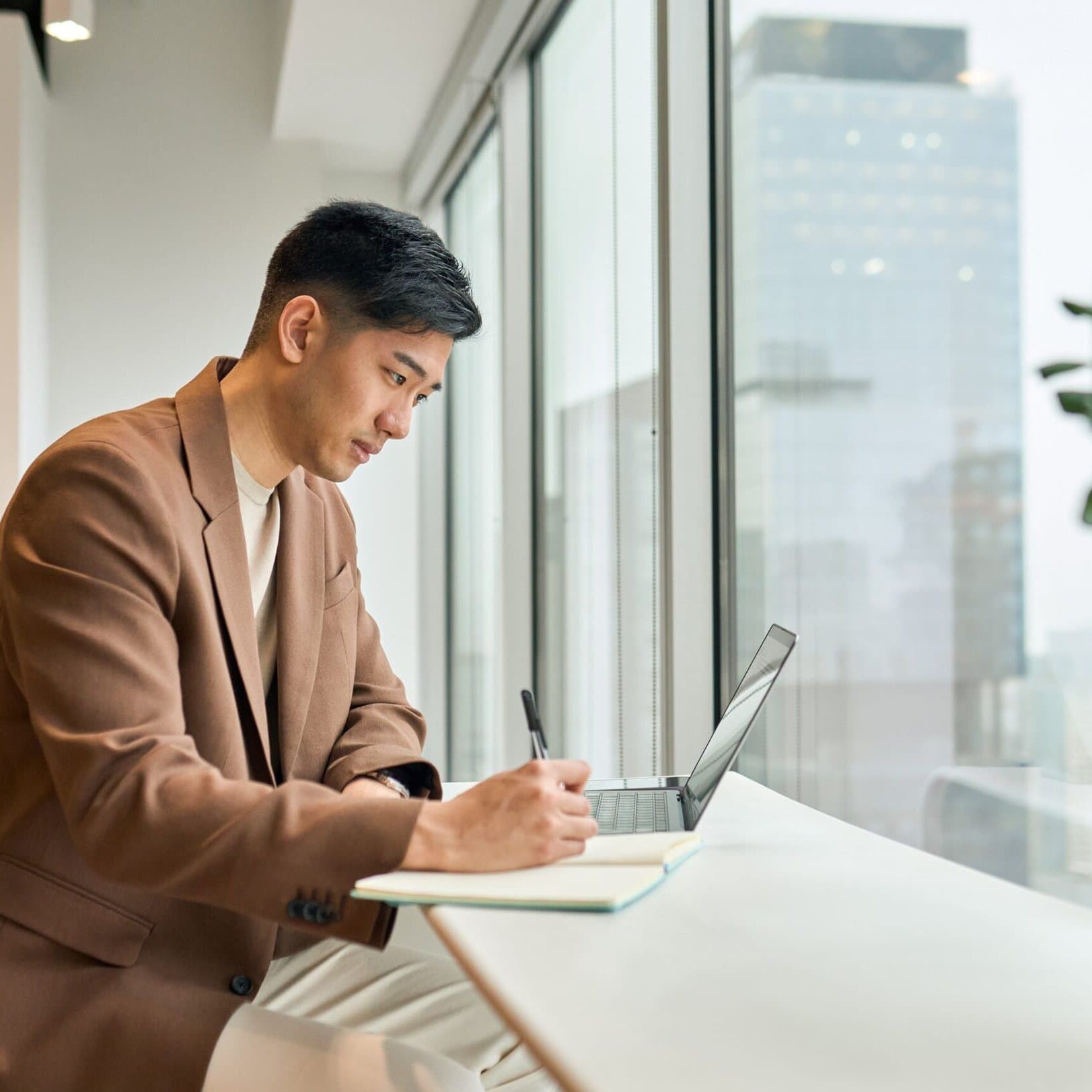 Young,Busy,Asian,Business,Man,Manager,Working,On,Laptop,In man sitting at table in front of window taking notes in a notebook while looking at a laptop